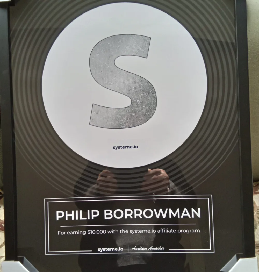 A framed systeme.io award. A large silver S sits inside a white circle on a black record-style disc, above a panel reading Philip Borrowman, for earning $10,000 with the systeme.io affiliate program, signed systeme.io and Aurelien Amacker.