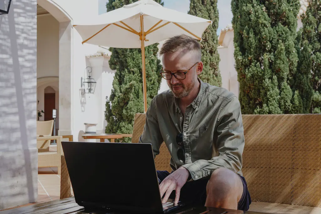 Philip Borrowman working on a laptop on a shaded outdoor terrace, sitting on a wicker sofa under a cream parasol, with cypress trees and a whitewashed building behind him.